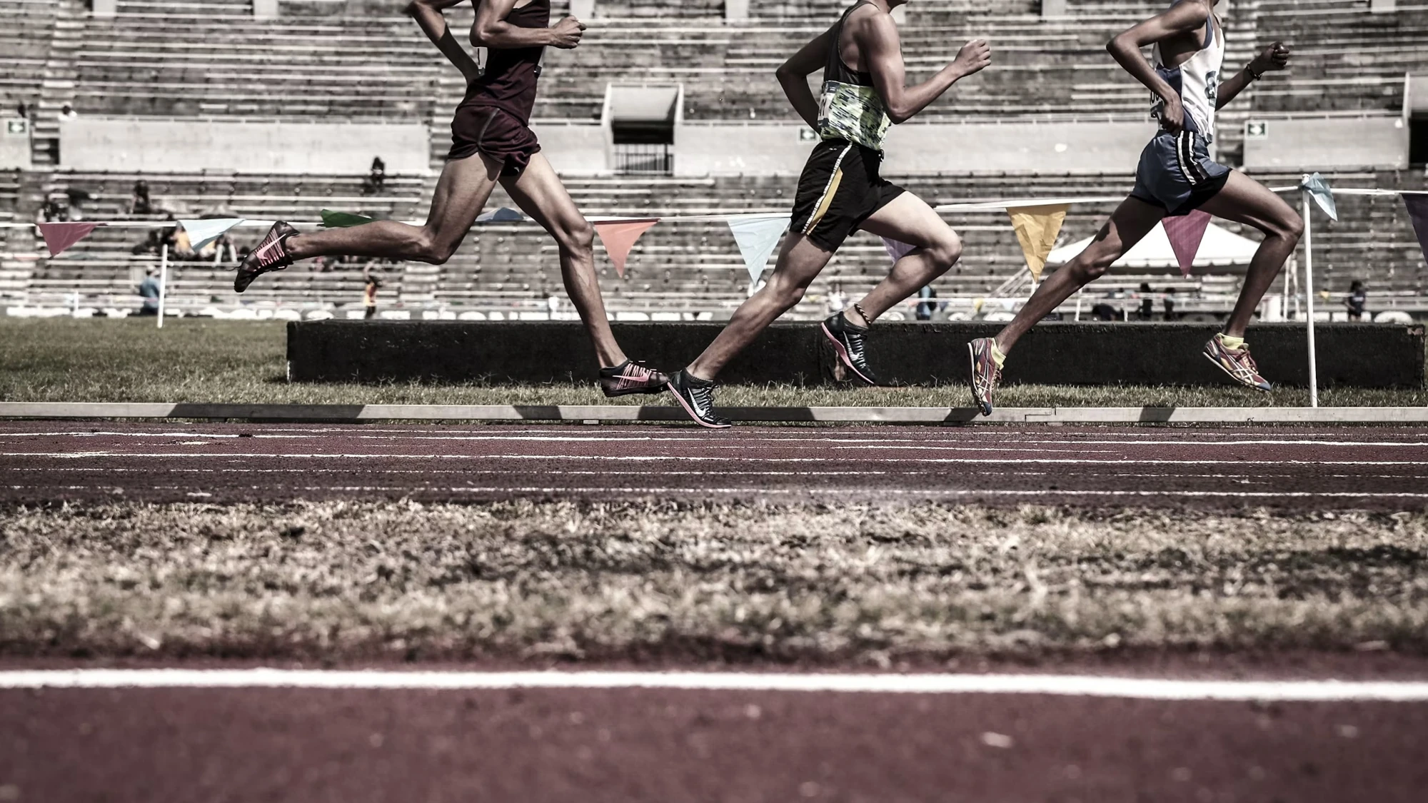 Three runners racing along an athletics track in front of stadium seating.